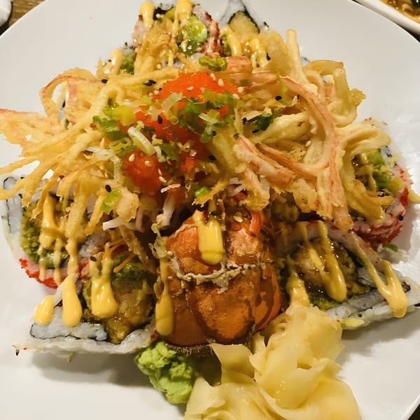 A large sushi roll topped with fried tempura, lobster, roe, and a creamy sauce.